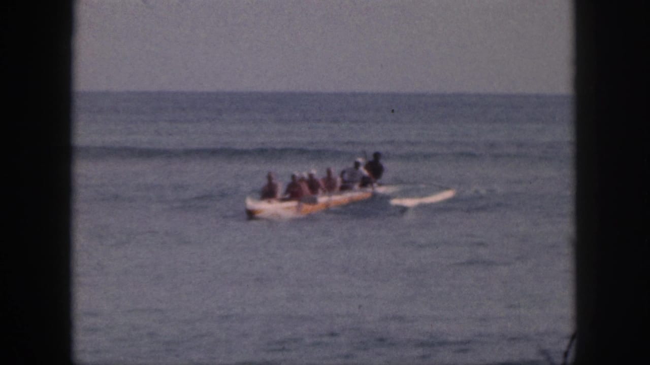 1959 Hawaii home-movie frame of a multi-person outrigger canoe moving across calm water under a hazy horizon.