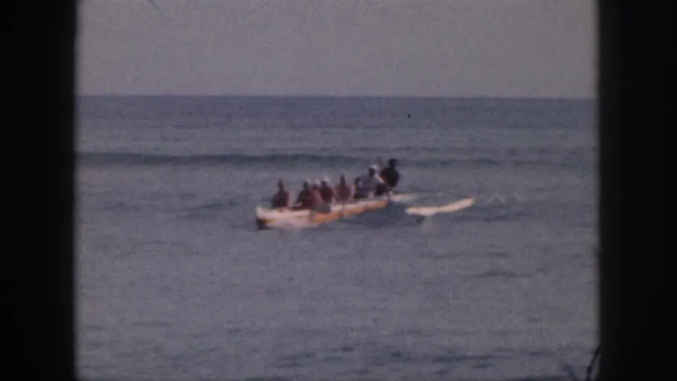 1959 Hawaii home-movie frame of a multi-person outrigger canoe moving across calm water under a hazy horizon.