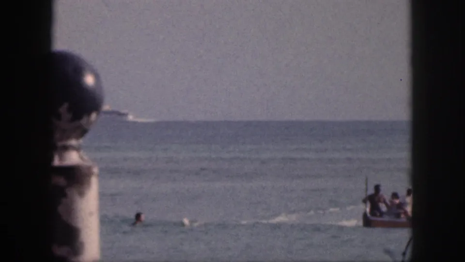 1959 Hawaii vacation footage frame with a boat partly entering the right edge, a swimmer, calm water, and shaded foreground framing.