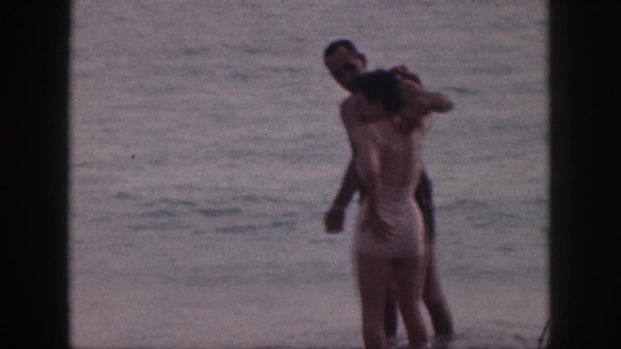 In 1959 Hawaii home-movie footage, a man and woman in swimwear embrace while standing in the surf.
