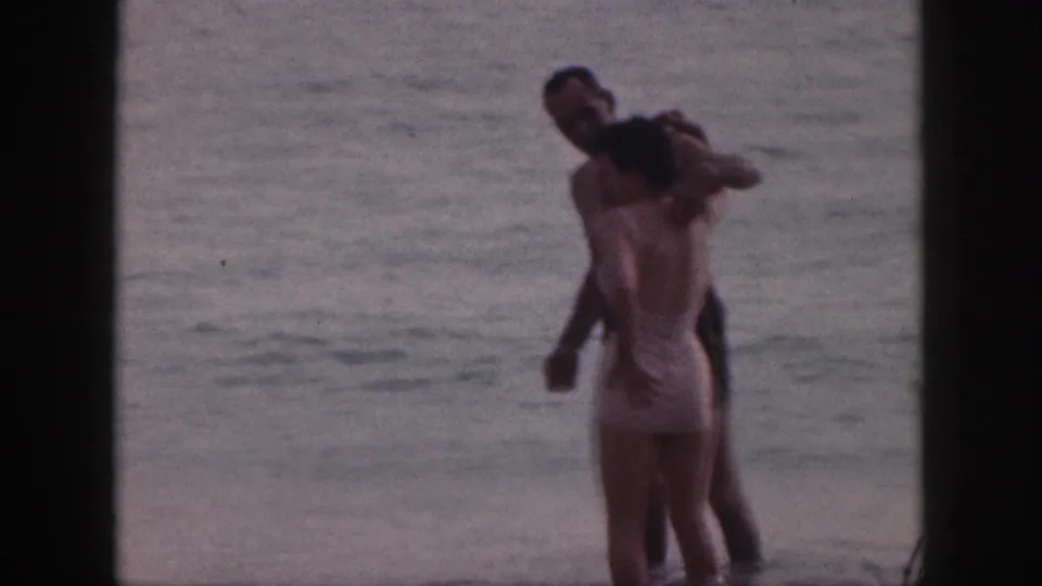 In 1959 Hawaii home-movie footage, a man and woman in swimwear embrace while standing in the surf.