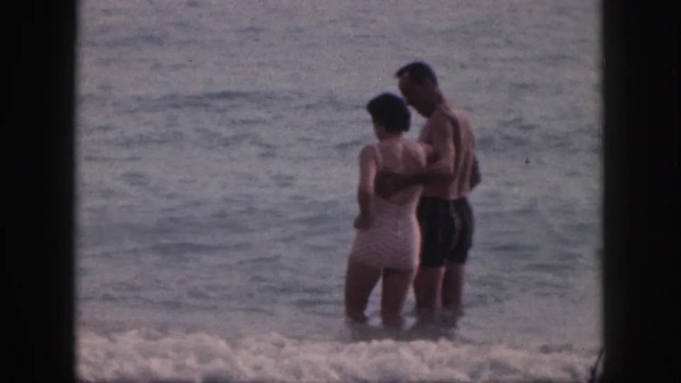 A couple stands waist-deep near breaking surf in 1959 Hawaii footage, with the man holding the woman at her back.