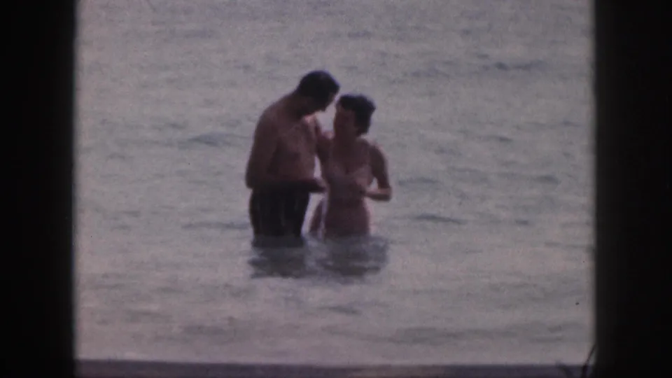 The man and woman stand close in waist-deep Hawaiian surf, captured in soft, grainy 8mm color footage.