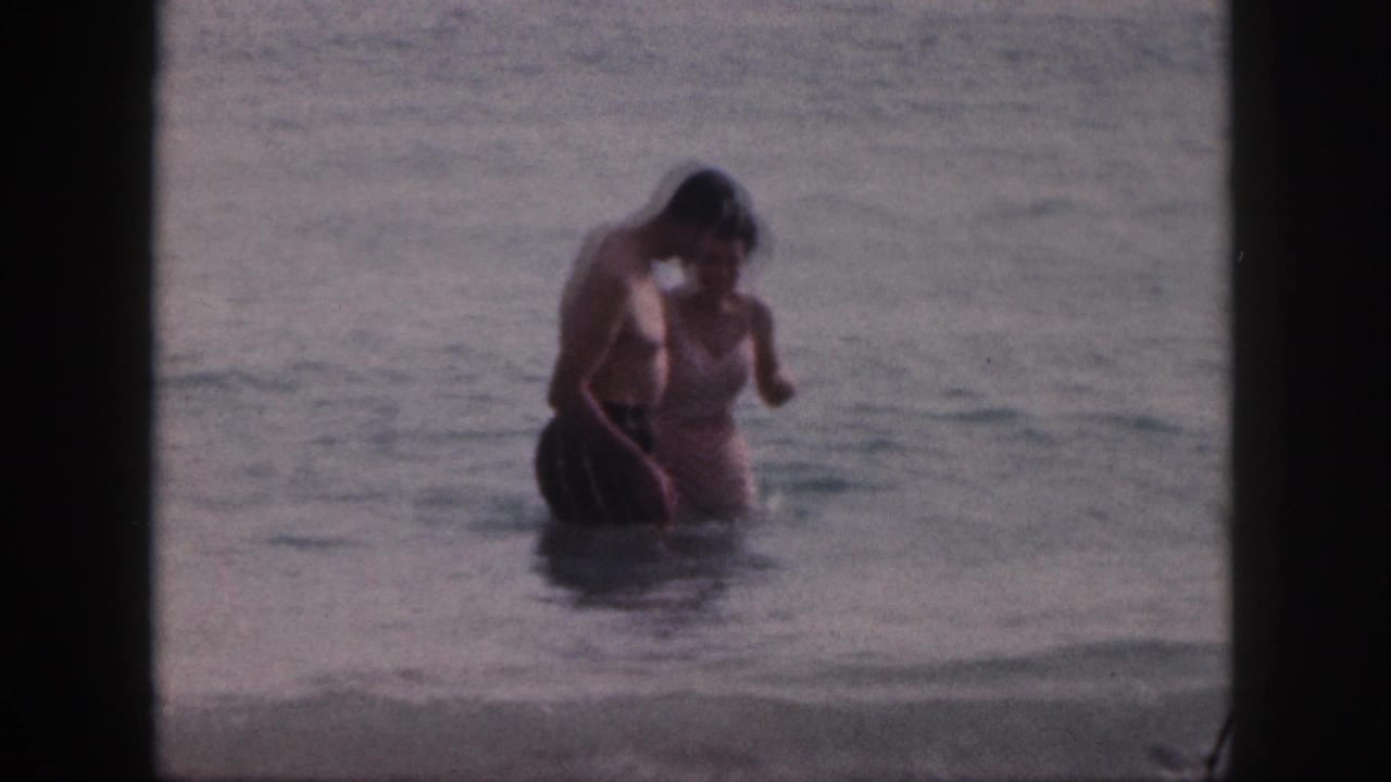 In 1959 Hawaii home-movie footage, an older couple stands close in shallow surf with dark frame borders around the scene.