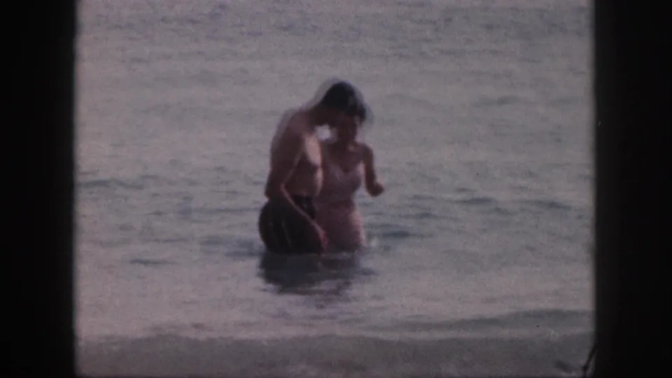 In 1959 Hawaii home-movie footage, an older couple stands close in shallow surf with dark frame borders around the scene.