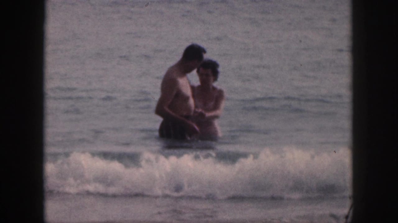 A couple stands in the surf as a small wave breaks in front of them.