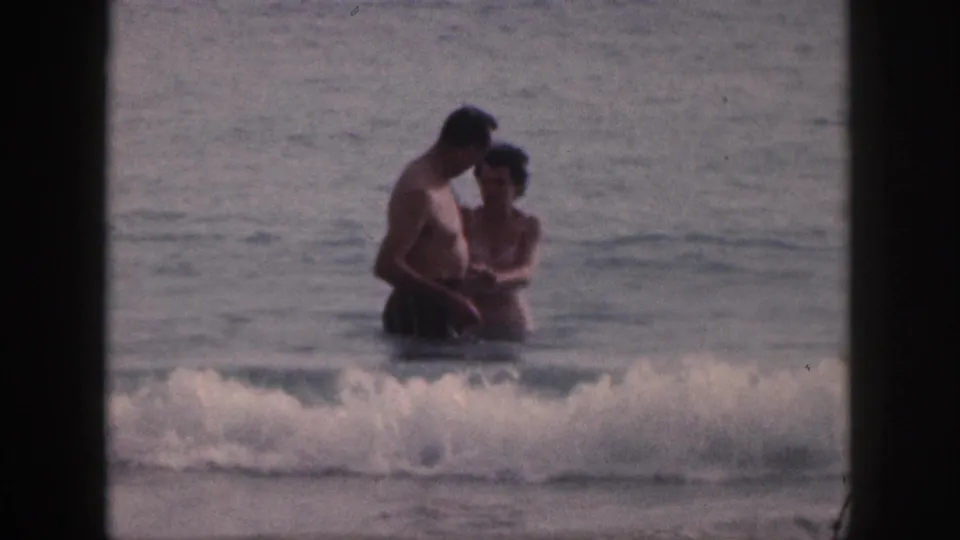 The couple remains close in shallow Hawaii surf, with a low wave crossing the foreground in soft 8mm color.