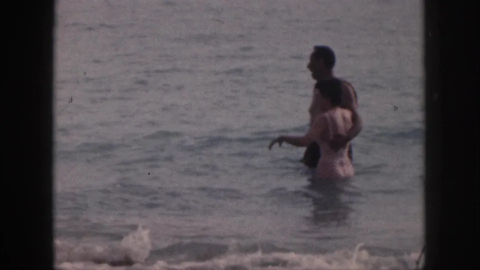 The couple wades in shallow water, the man in dark trunks and the woman in a light swimsuit, seen in soft home-movie focus.