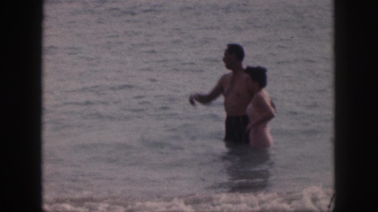 In 1959 Hawaii home-movie footage, a man gestures while standing beside a woman in shallow surf.
