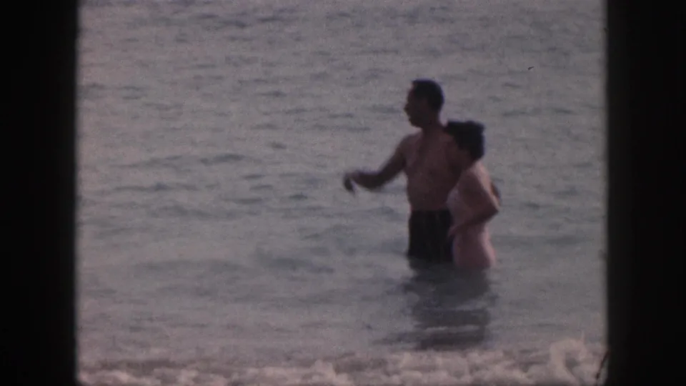 In 1959 Hawaii home-movie footage, a man gestures while standing beside a woman in shallow surf.