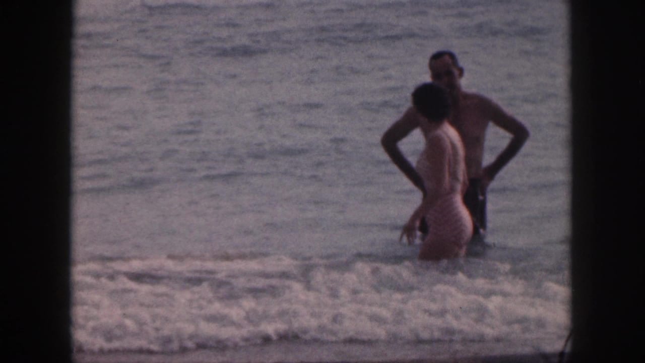 A man and woman in swimsuits stand in shallow Hawaii surf, framed by dark 8mm film borders.