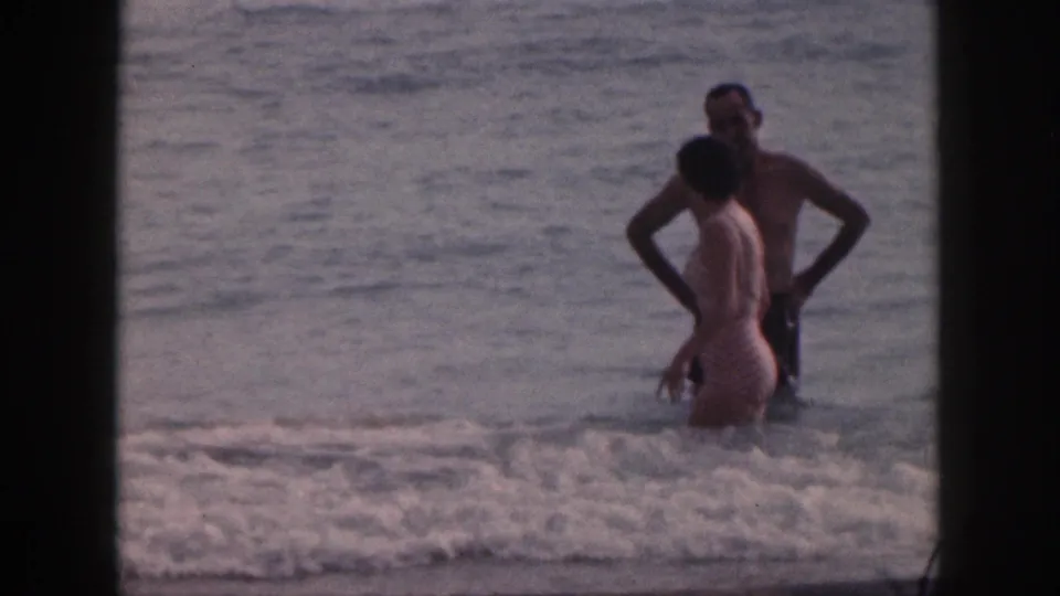 A man and woman in swimsuits stand in shallow Hawaii surf, framed by dark 8mm film borders.