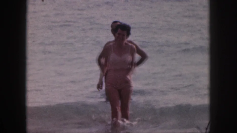 A woman in a light patterned swimsuit walks toward shore while a man stands behind her in the water.