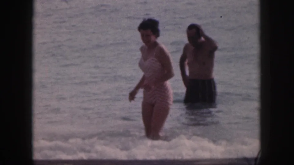 In the closing frame, the woman moves through small surf with the man standing farther back in the water.