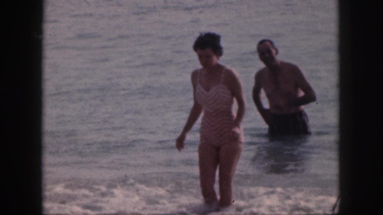 In 1959 Hawaii home-movie footage, a woman in a striped one-piece swimsuit walks through shallow ocean water with a shirtless man behind her.