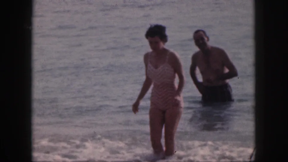 In 1959 Hawaii home-movie footage, a woman in a striped one-piece swimsuit walks through shallow ocean water with a shirtless man behind her.