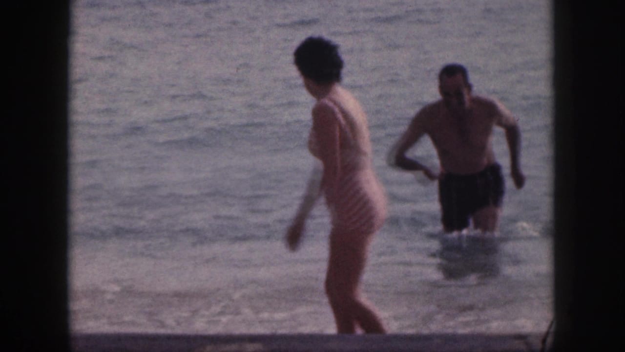 Woman turns in shallow water as a shirtless man moves through the surf behind her.