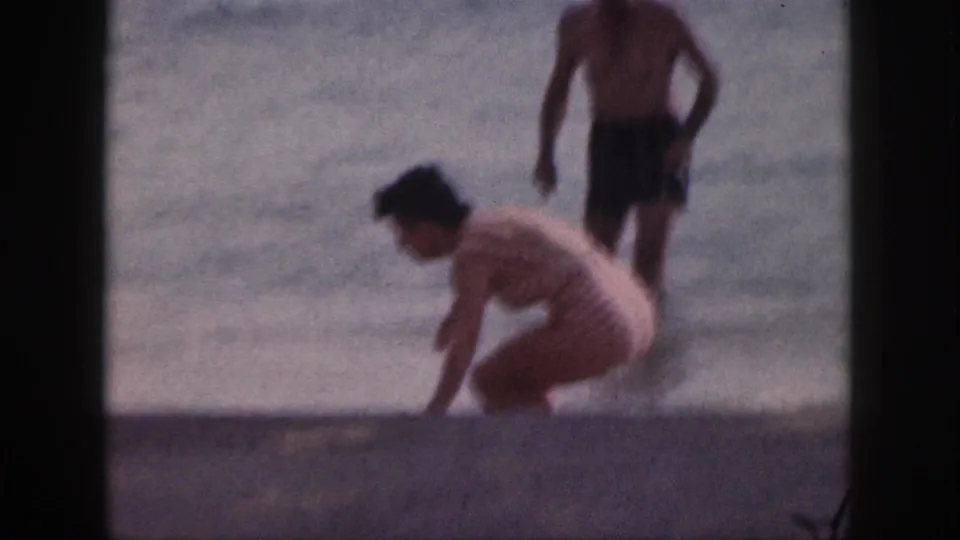 The woman crouches at the edge of the water in the 1959 Hawaii beach footage, with the man standing farther back in soft focus.