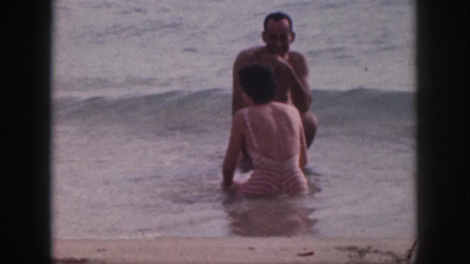 The couple faces each other in shallow surf in the Hawaii home movie, with the woman seated or crouched and the man smiling toward her.