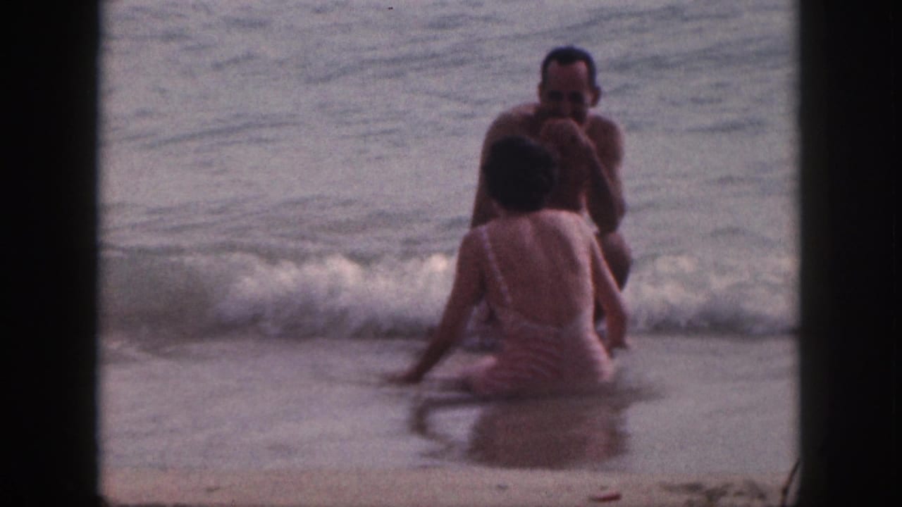 Two adults sit in shallow ocean surf as small waves roll onto the beach.