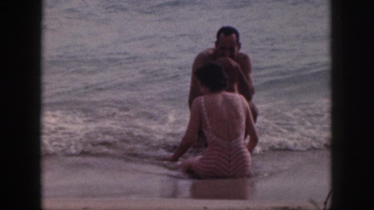 A seated couple faces each other in the surf near a breaking wave.