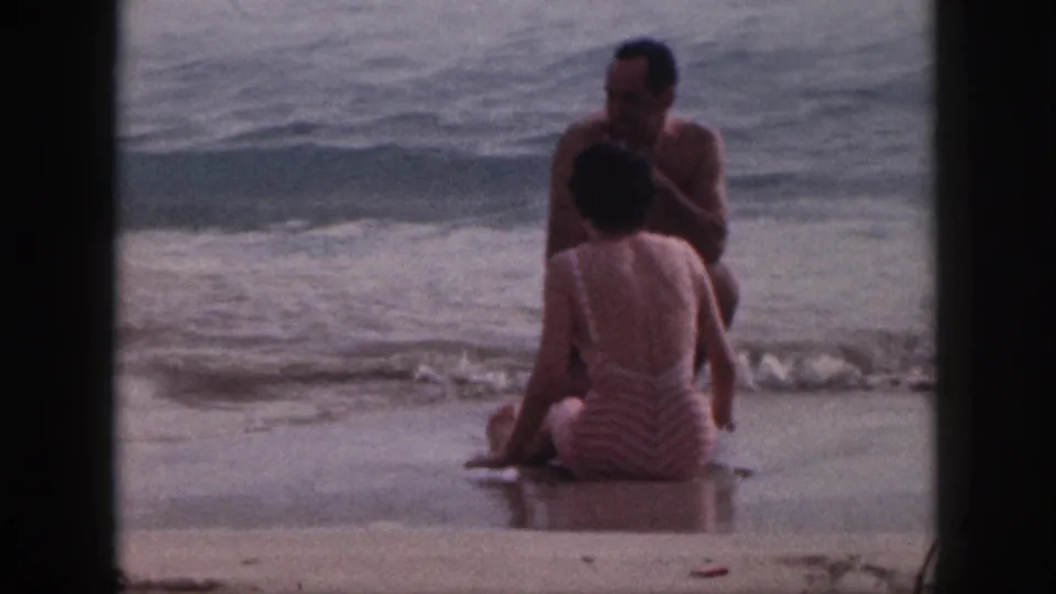 A 1959 Hawaii 8mm frame shows the seated beach couple from behind and front, with muted color, grain, and surf rolling in.