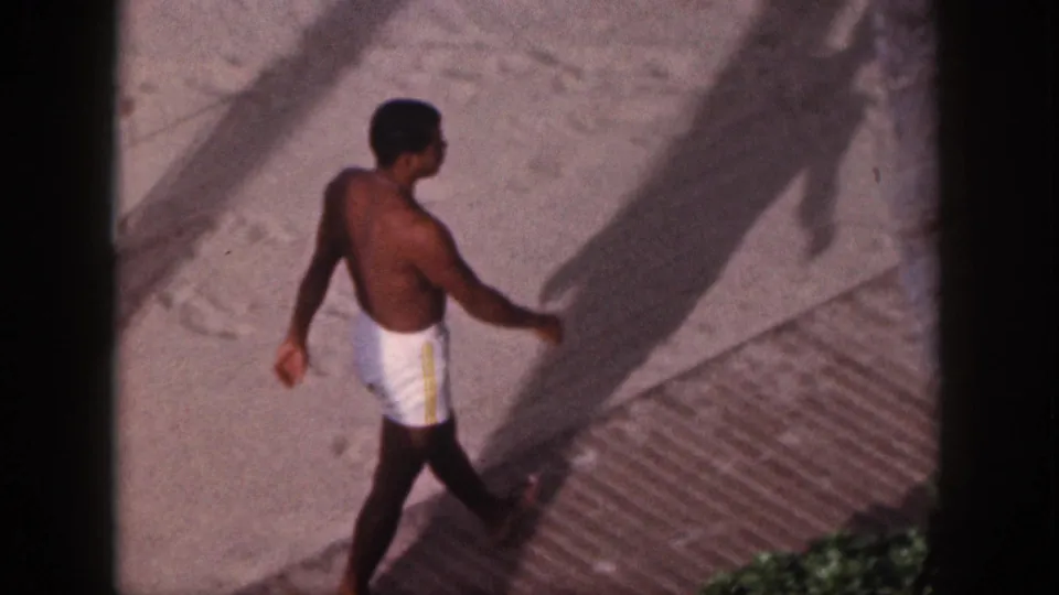 1959 Hawaii beach-vacation frame showing a man in white swim trunks walking along sand beside a wooden boardwalk and palm shadow.