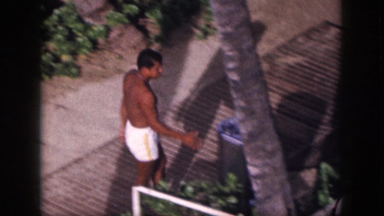 1959 Hawaii home-movie frame with a man in white swim trunks nearing a water fountain along a shaded tropical walkway.