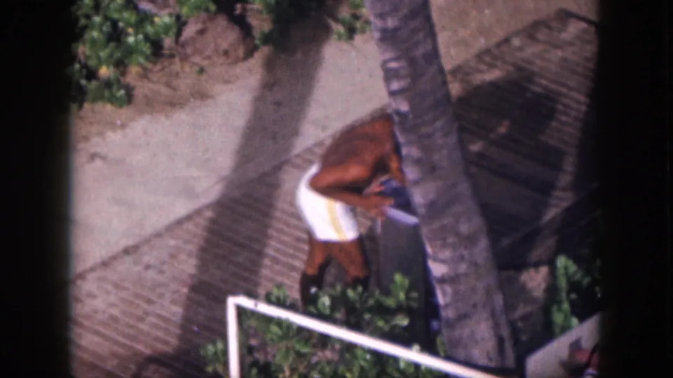 1959 Hawaii home-movie frame showing the man bending to drink from an outdoor fountain beside tropical planting.