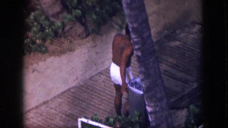 1959 Hawaii home-movie frame of a shirtless man in white swim trunks walking barefoot past palms and a trash can on a resort path.