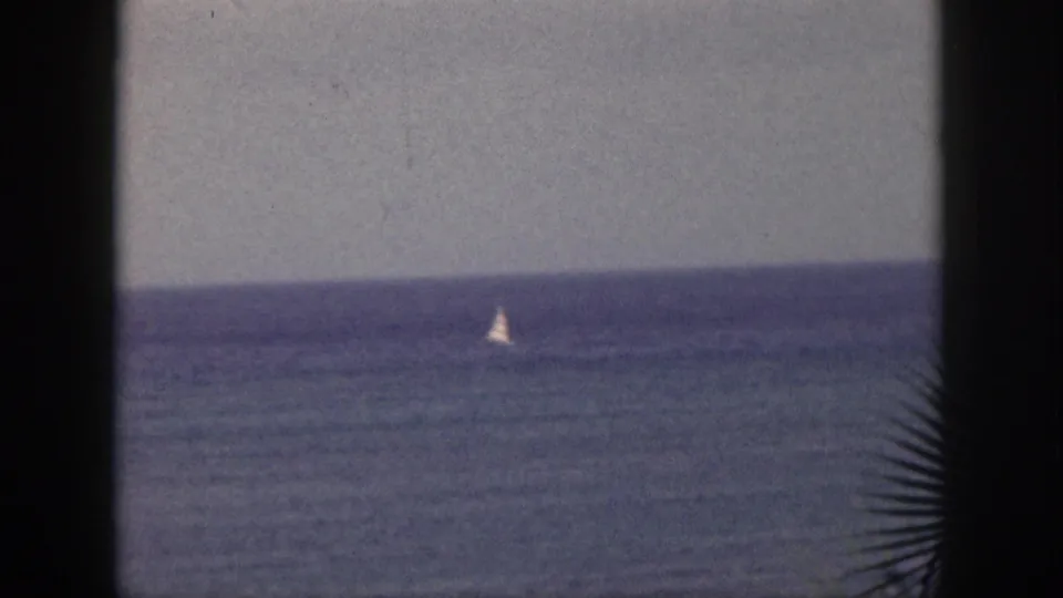 1959 Hawaii 8mm frame showing a small white sailboat far out on the ocean, seen through dark foreground edges under a gray sky.