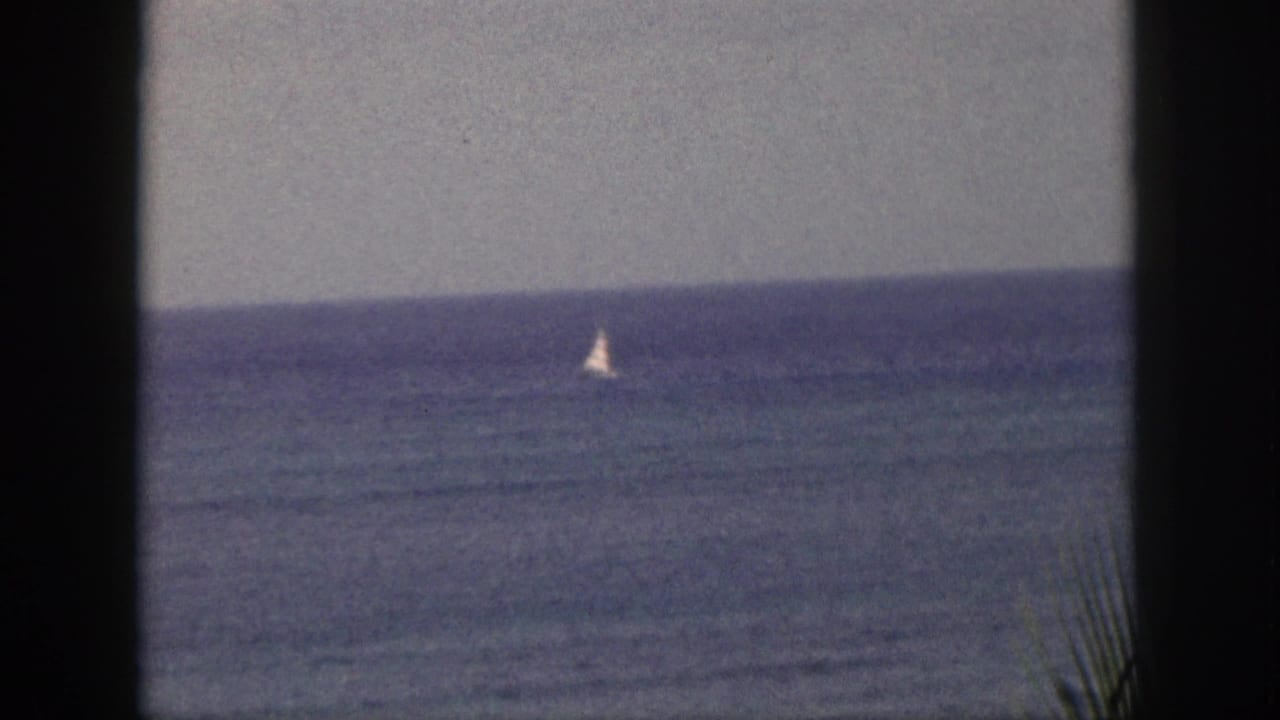 A small sailboat appears on the 1959 Hawaii ocean horizon, viewed through dark foreground framing on 8mm film.