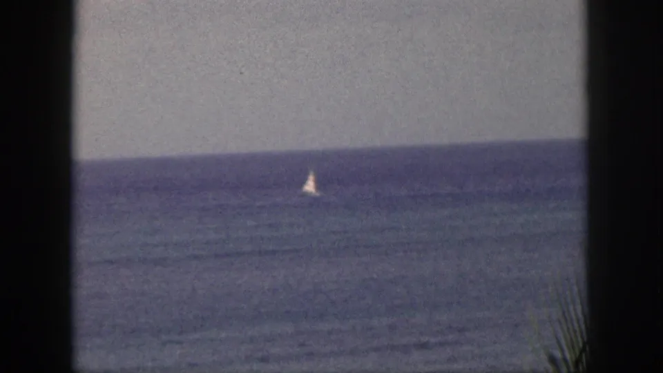 A small sailboat appears on the 1959 Hawaii ocean horizon, viewed through dark foreground framing on 8mm film.