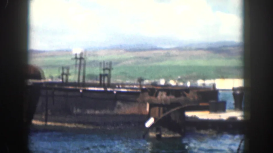 1959 Hawaii 8mm frame looking past a dark pier structure across water toward green hills under clouds.