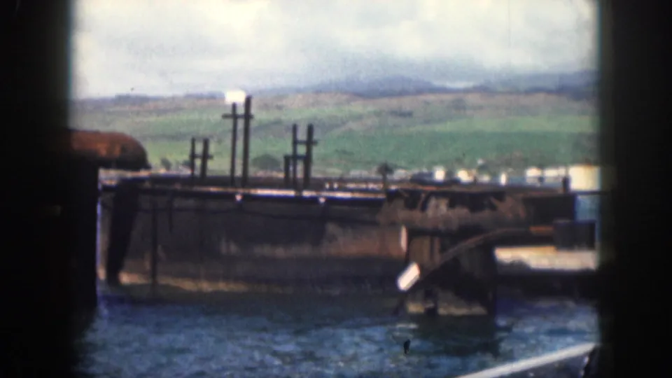 1959 Hawaii 8mm frame of a weathered waterfront structure with water, distant shoreline, and green hills.