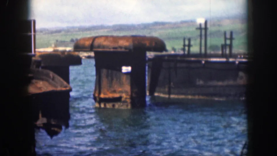 Final 8mm frame from Hawaii showing weathered waterfront concrete forms, upright posts, blue water, and hills under a cloudy sky.