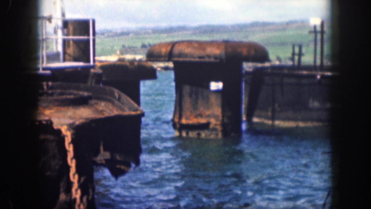 1959 Hawaii home-movie frame showing blue water, weathered pier supports, a platform, and green hills in the distance.
