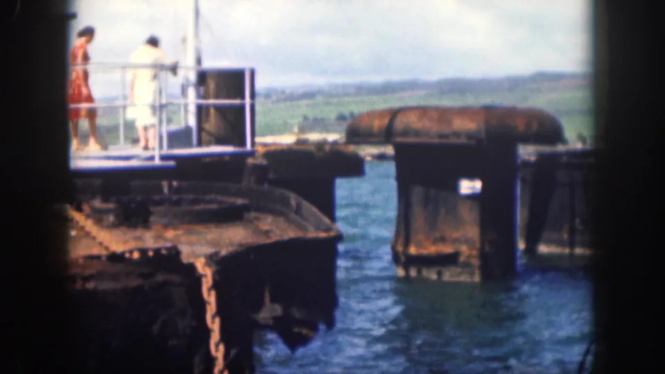 1959 Hawaii waterfront scene with two adults on a metal pier platform, blue water, rusted supports, and distant hills.