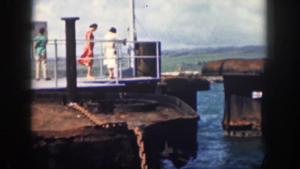 1959 Hawaii home-movie frame showing three adults around a railed waterfront platform with water and distant hills behind them.
