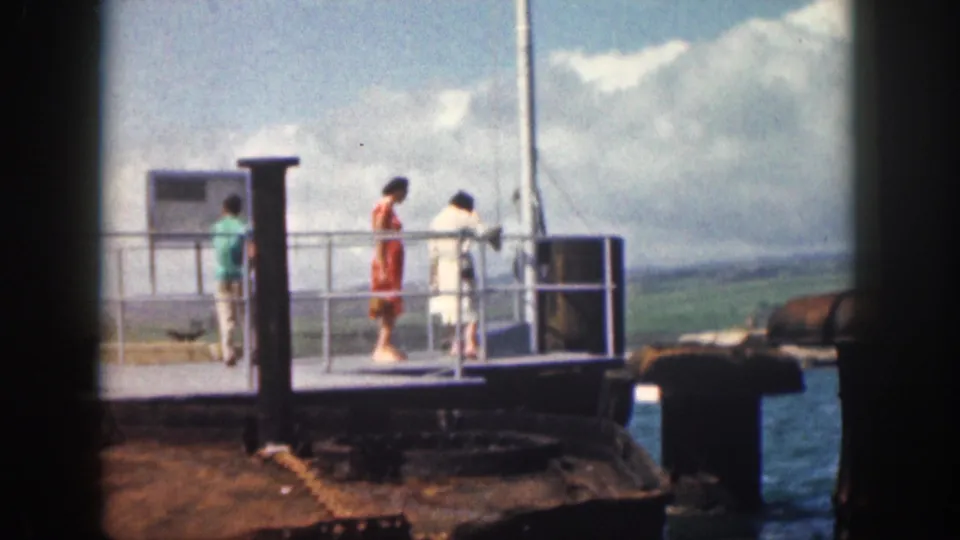 1959 Hawaii waterfront frame with three adults on a metal platform beside blue water, a cloudy sky, and green hills.