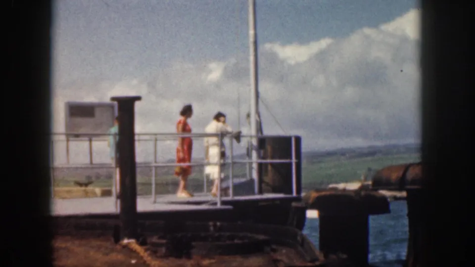1959 Hawaii home-movie frame showing people at a naval station dock railing, including a woman in red and a person in white, looking over the water.