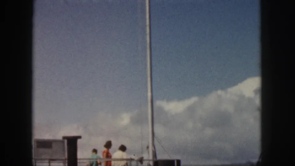 1959 Hawaii frame with visitors near a waterfront railing, a tall flagpole, harbor water, and large clouds in the background.