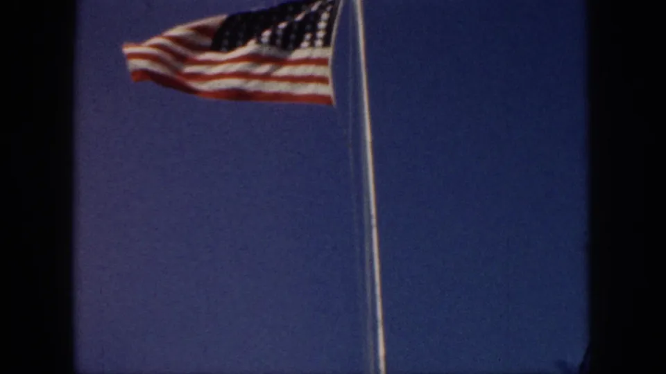 1959 Hawaii home-movie frame focused on a U.S. flag waving from a tall pole against a bright blue sky.