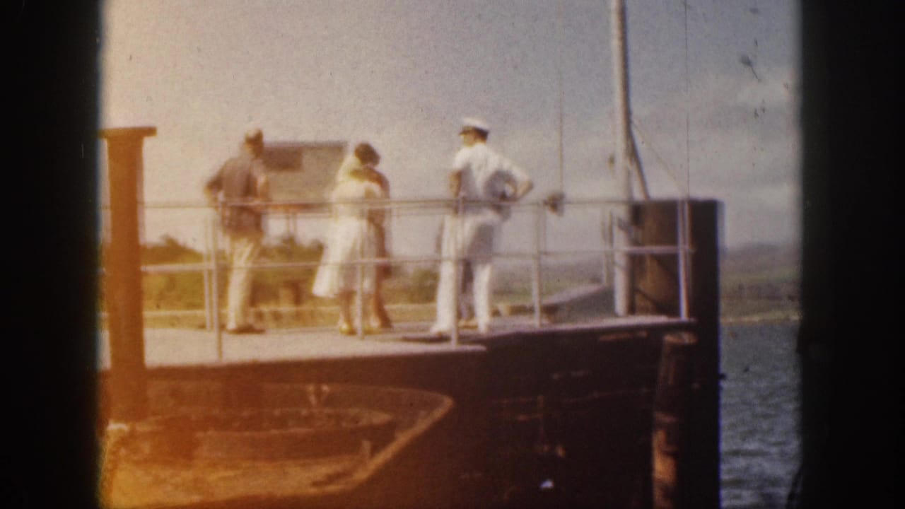 A few people stand along a railing on a dock or vessel, with a sailor at the right.