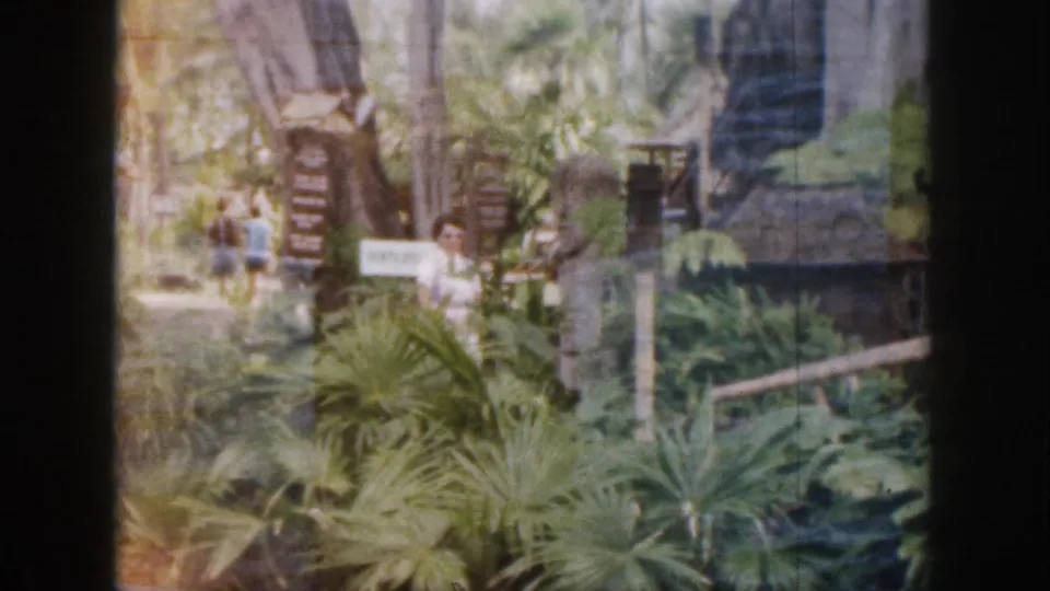1959 Hawaii 8mm frame of people walking through a leafy tropical garden with wooden signs and rustic buildings nearby.