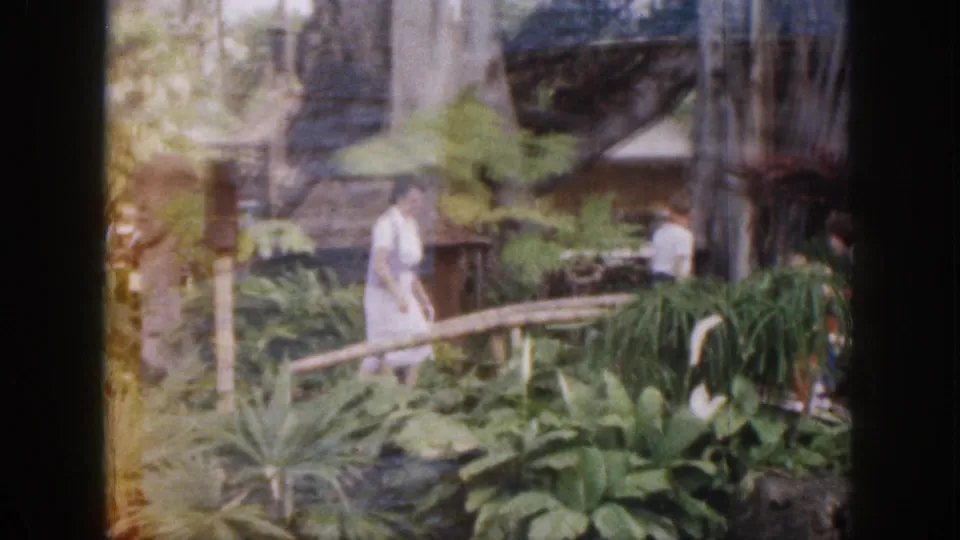 1959 Hawaii home-movie still showing a woman in a white dress near a rail, surrounded by broad-leaf plants and rustic garden structures.