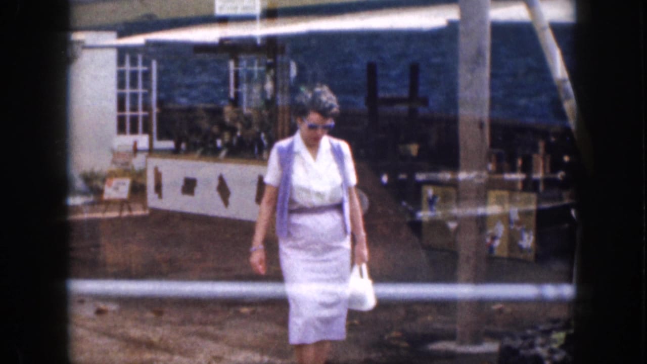 Woman in a white dress and sunglasses walks past low buildings and outdoor seating.
