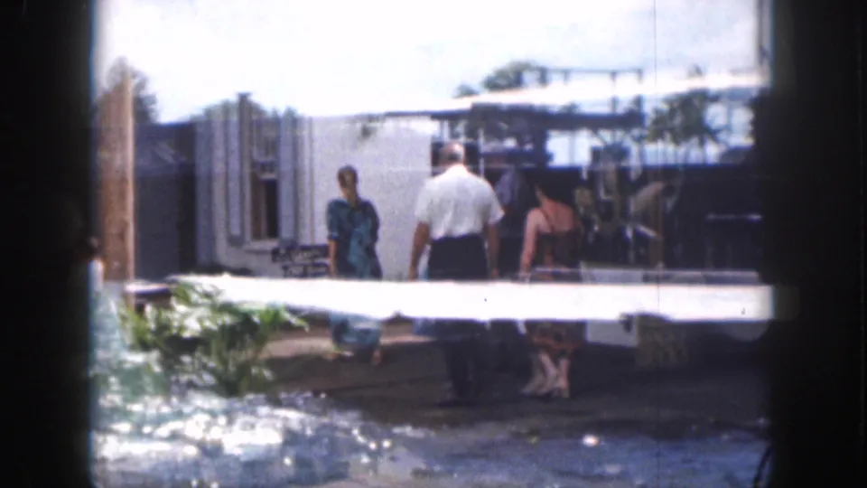 Adults walk away through a bright outdoor public area in 1959 Hawaii, with low buildings, plants, and shaded seating in view.