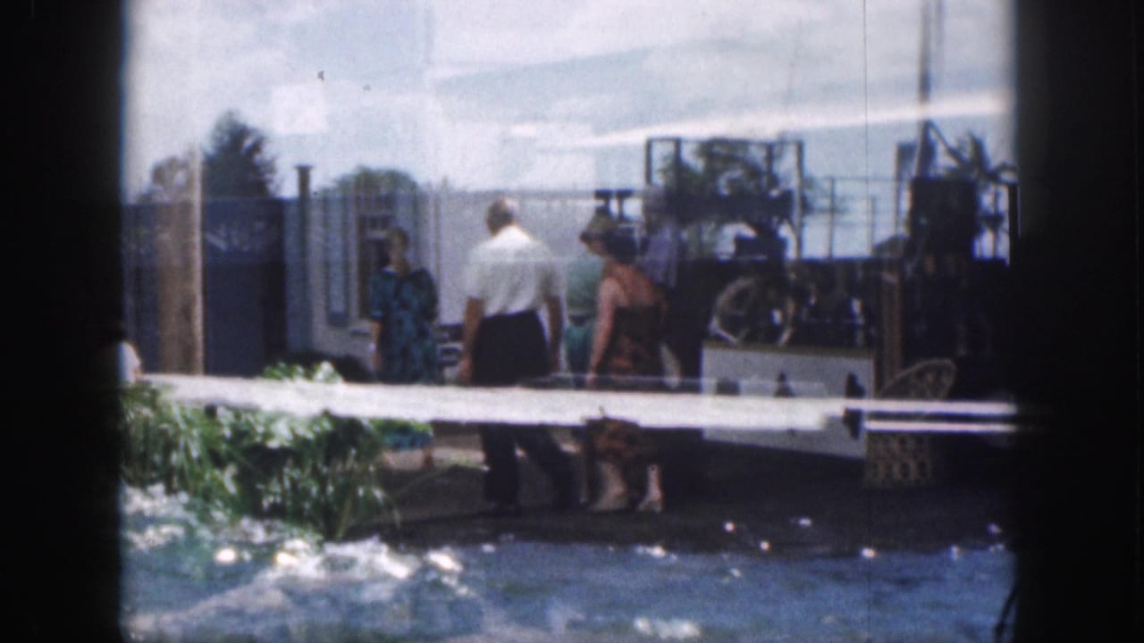 Adults walk along a waterfront pier with water in the foreground and resort structures behind them.