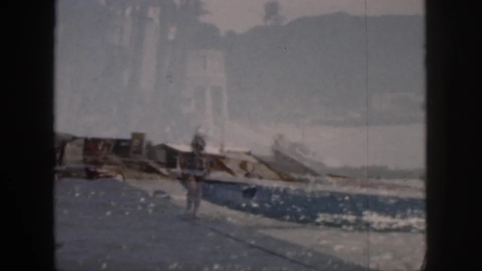 1959 Hawaii frame of a man moving along the promenade as small boats sit near the bright, hazy waterfront.